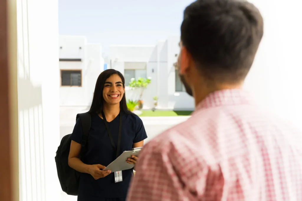 Clinician with a backpack and tablet greeting a parent at the front door of a home