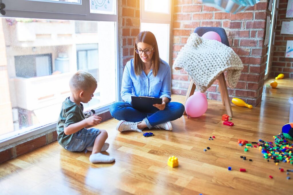 Therapist taking notes while a young boy plays with toys on the floor at home