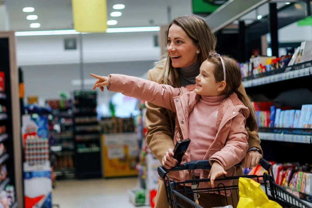 Mother pushing her young daughter in a shopping cart through a grocery store aisle
