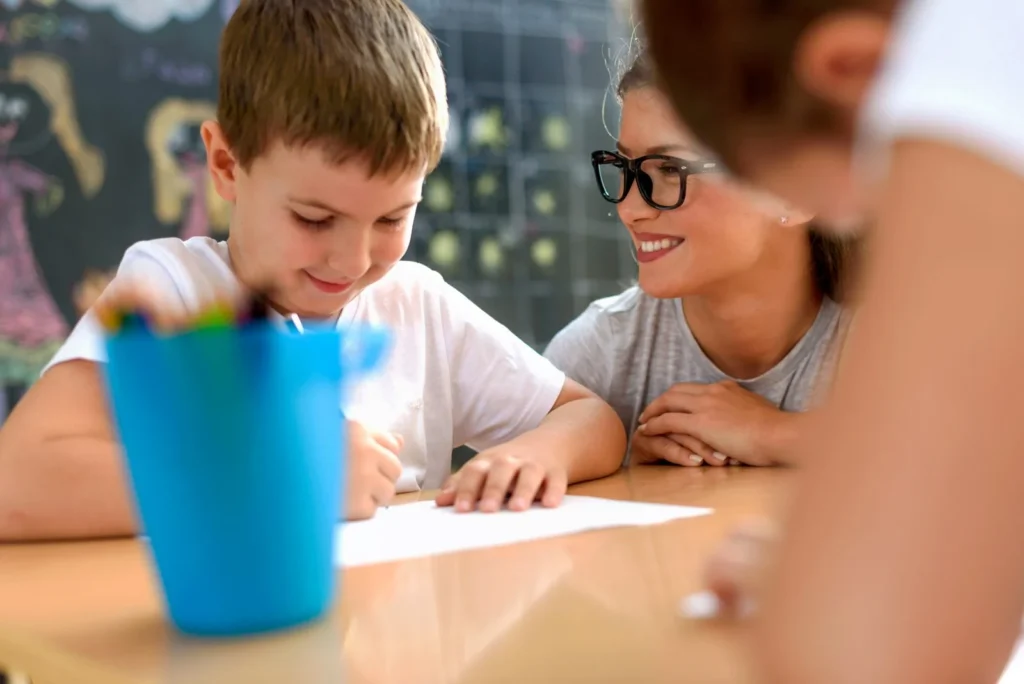 Teacher smiling at a young boy as he writes at his desk