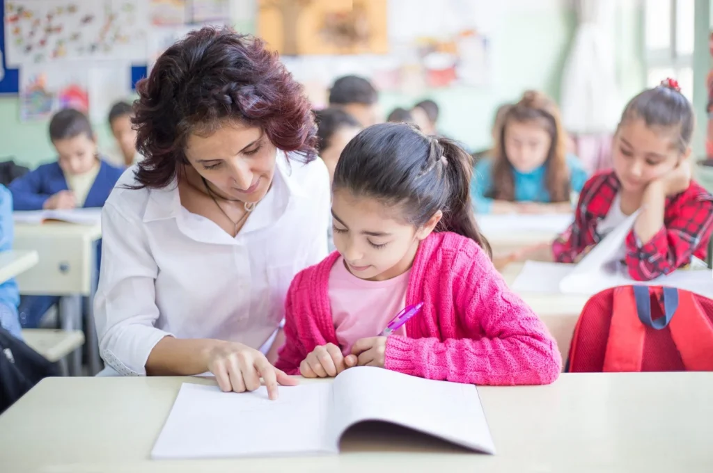 Teacher guiding a young girl through her work in a busy classroom