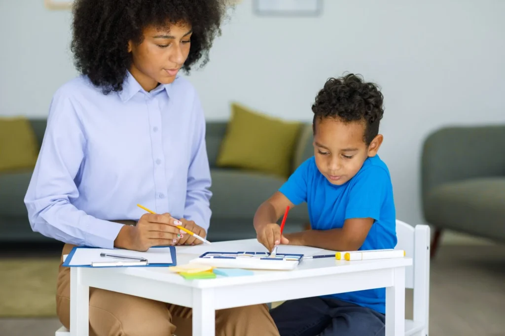 Therapist taking notes while a young boy colors at a small table at home