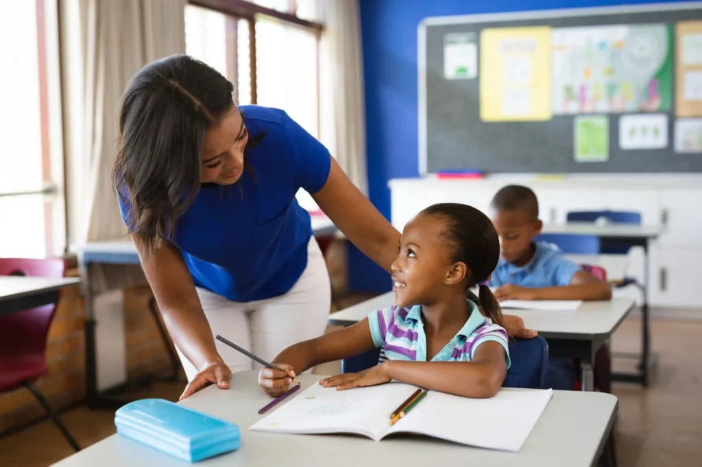 Teacher leaning over to help a smiling girl with her classwork