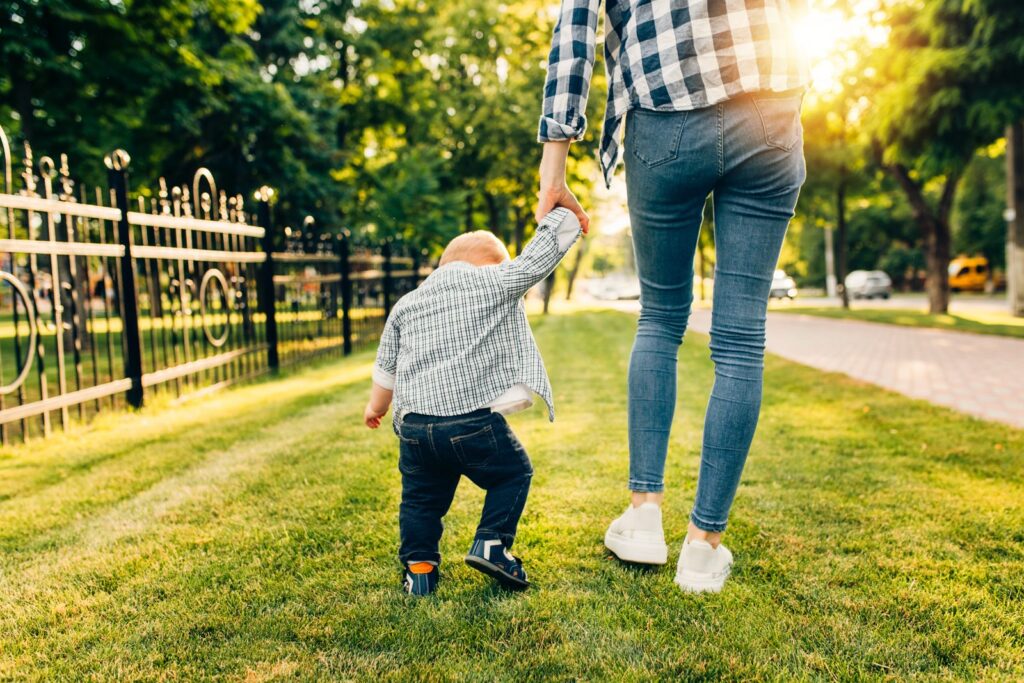 Toddler holding his mother’s hand while walking through a park