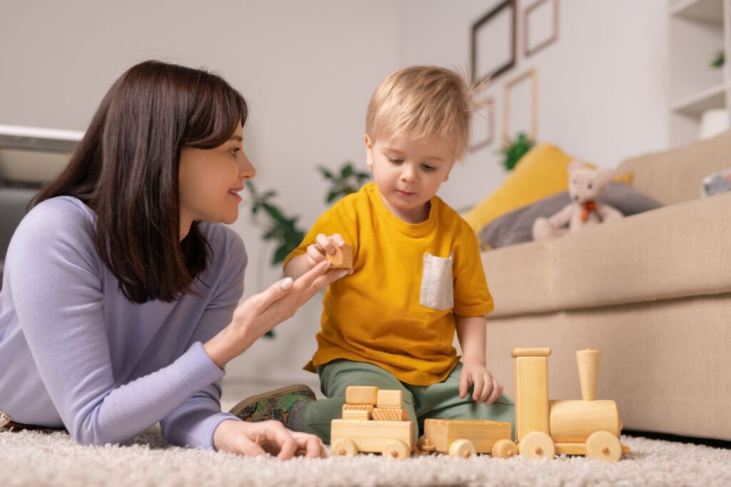 Mother teaching her toddler son to stack wooden blocks on the living room carpet